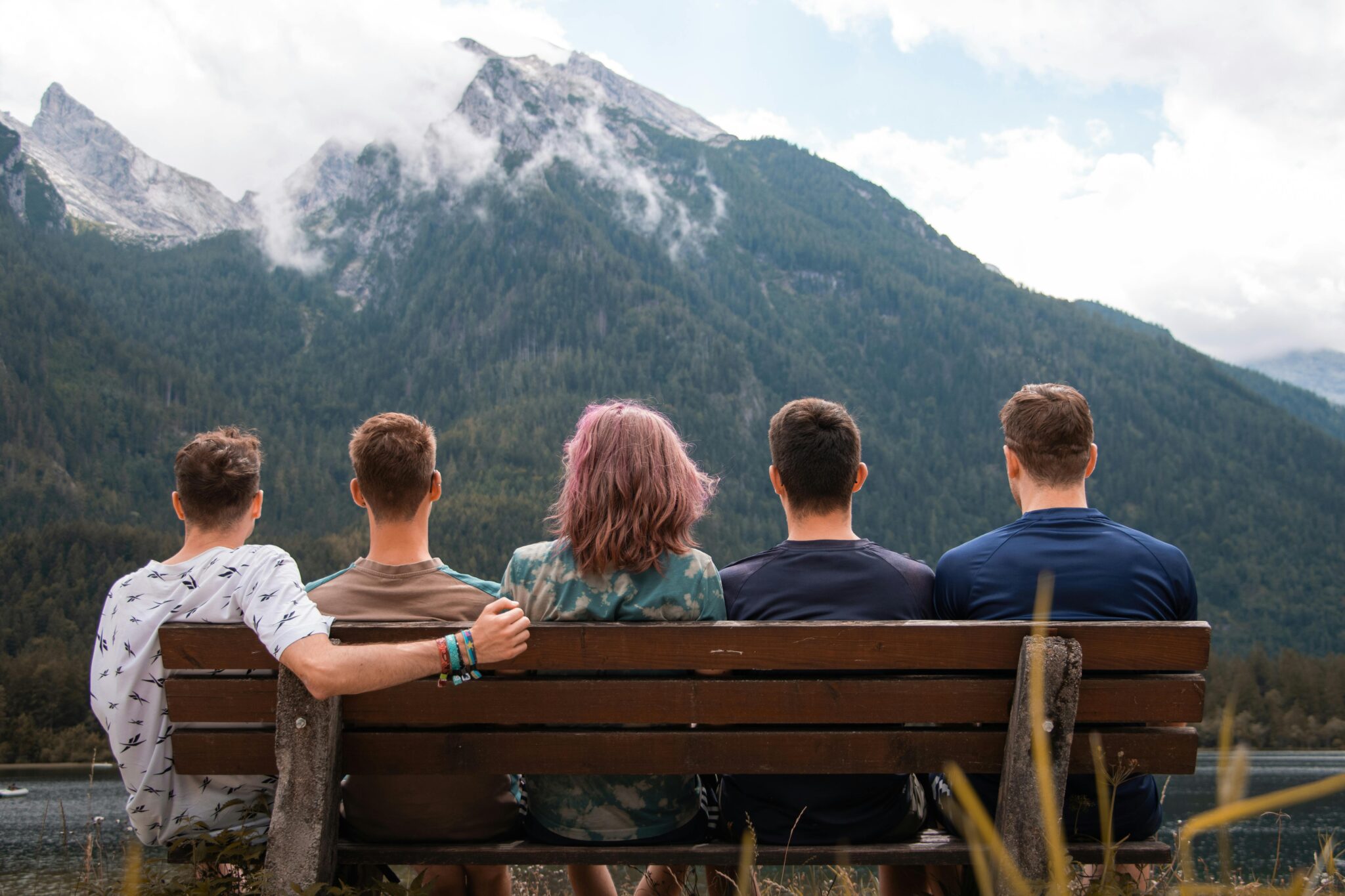 Un groupe de cinq jeunes vus de dos, assis ensemble sur un banc en bois face à un paysage de montagnes et un lac.
